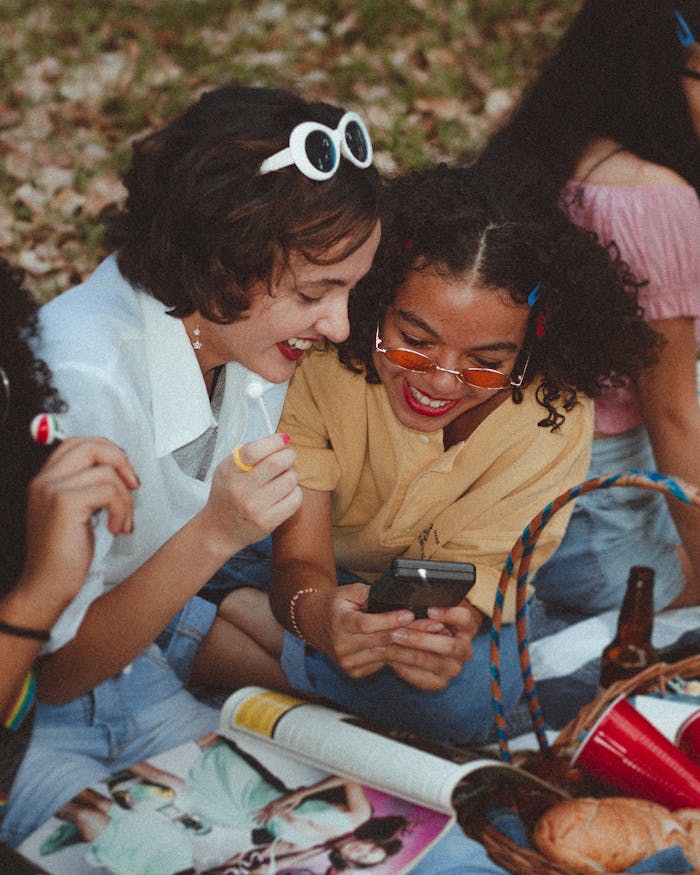 gallery-03 A group of happy young women enjoying a sunny picnic outdoors, engaging with a smartphone and laughing.