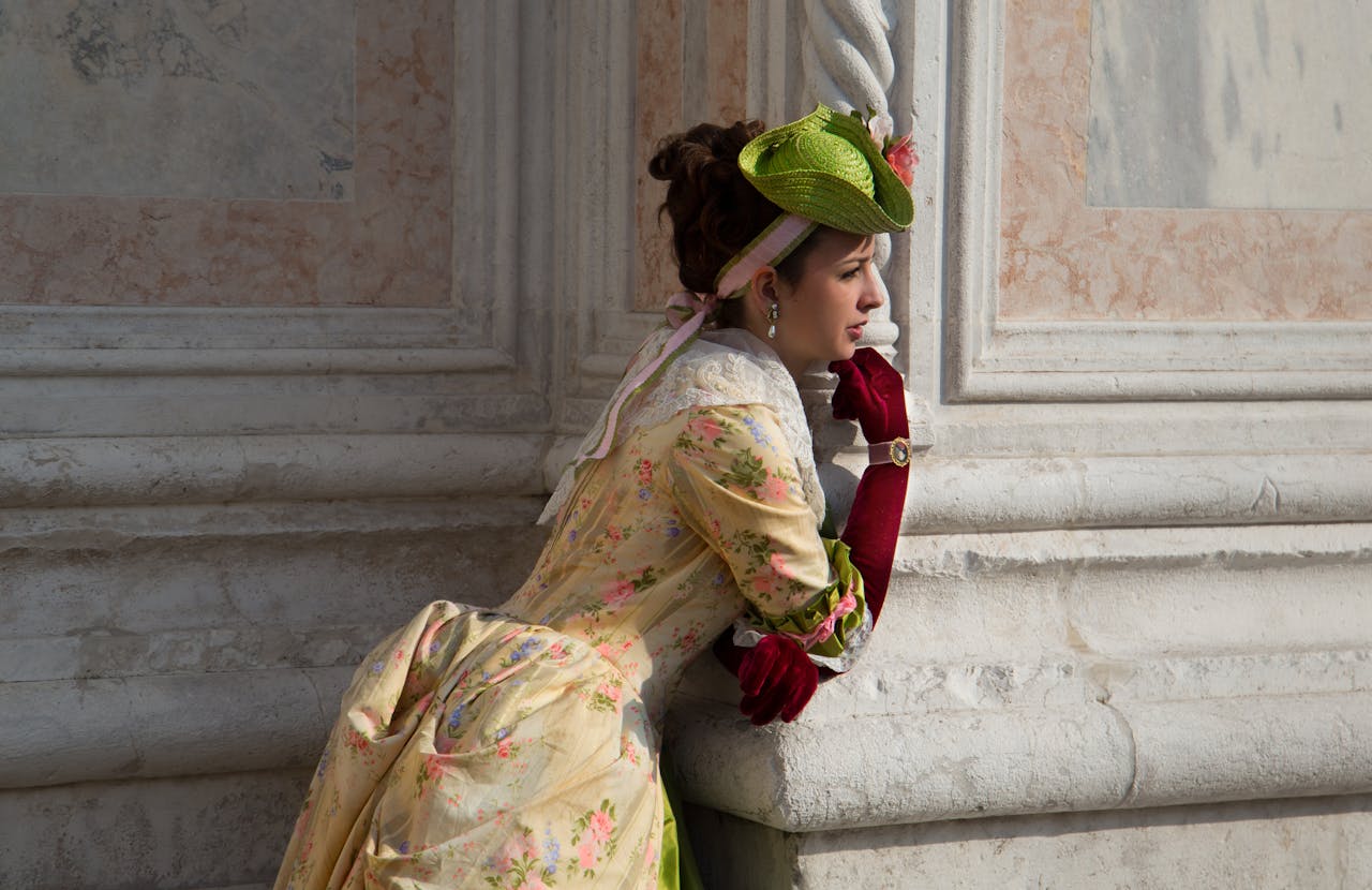 gallery-01 Elegant woman in vintage dress with a green hat, leaning on stone architecture outdoors.