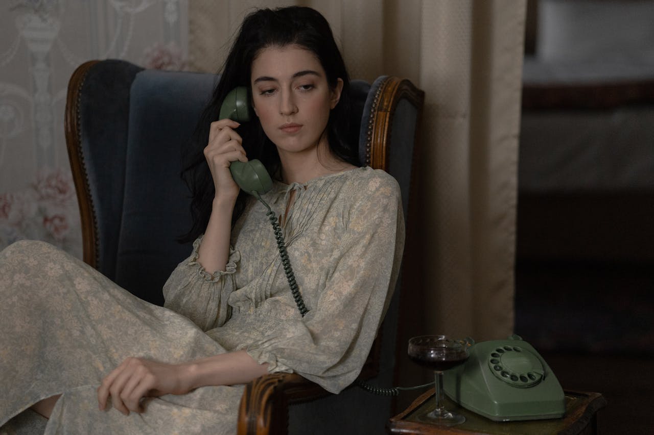 footer-cta-img Woman in vintage dress sitting in a chair, talking on an old-fashioned phone indoors.